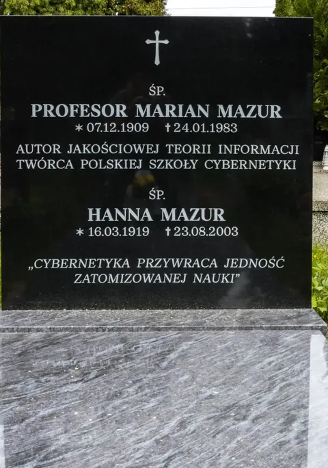 Colour photograph: a black granite headstone inscribed “Profesor Marian Mazur, autor jakościowej teorii informacji, twórca Polskiej Szkoły Cybernetyki” (Professor Marian Mazur, author of the qualitative theory of information, founder of the Polish School of Cybernetics) with the quotation “Cybernetics restores the unity of atomised science”.